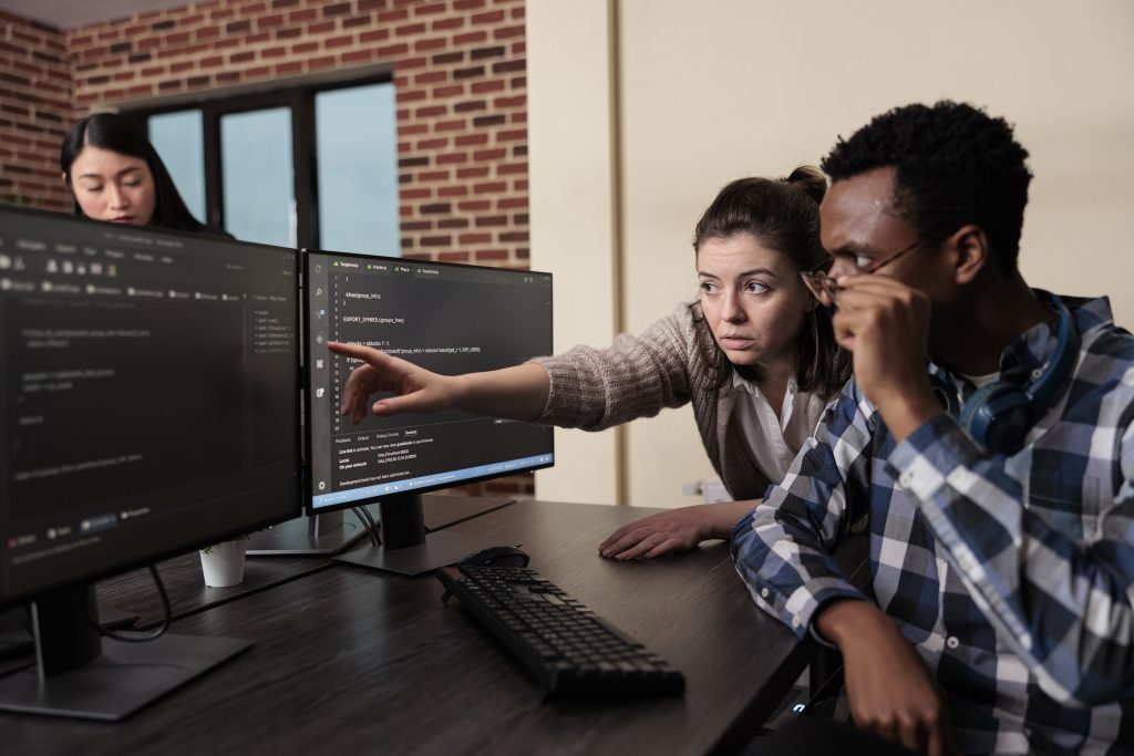 caucasian-software-developer-signaling-screen-data-breach-deactivated-security-system-tech-engineer-observing-multiple-system-security-breaches-caused-by-overloaded-storage-servers - Vantree Two coworkers review code on dual computer monitors as one points at the screen during a collaborative debugging session in an office with exposed brick wall (informative, concise).