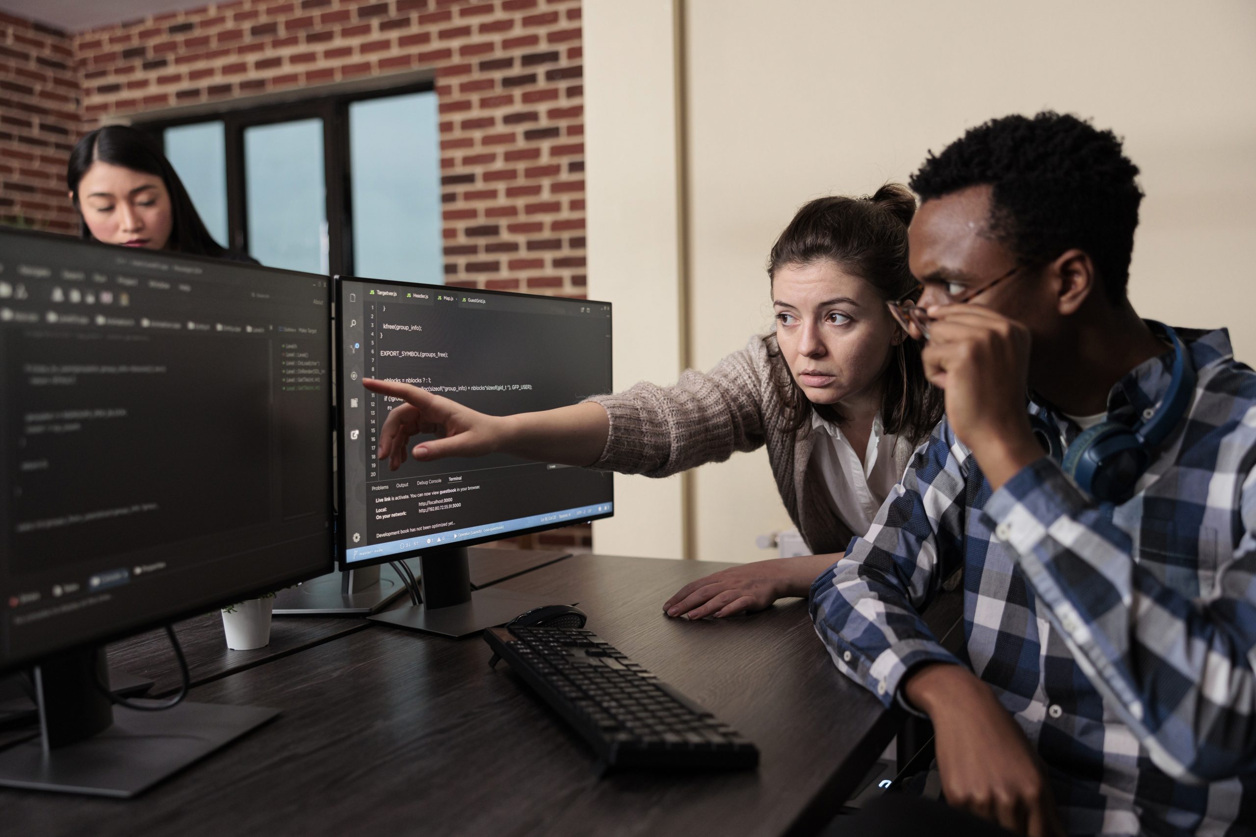 Two coworkers review code on dual computer monitors as one points at the screen during a collaborative debugging session in an office with exposed brick wall (informative, concise).