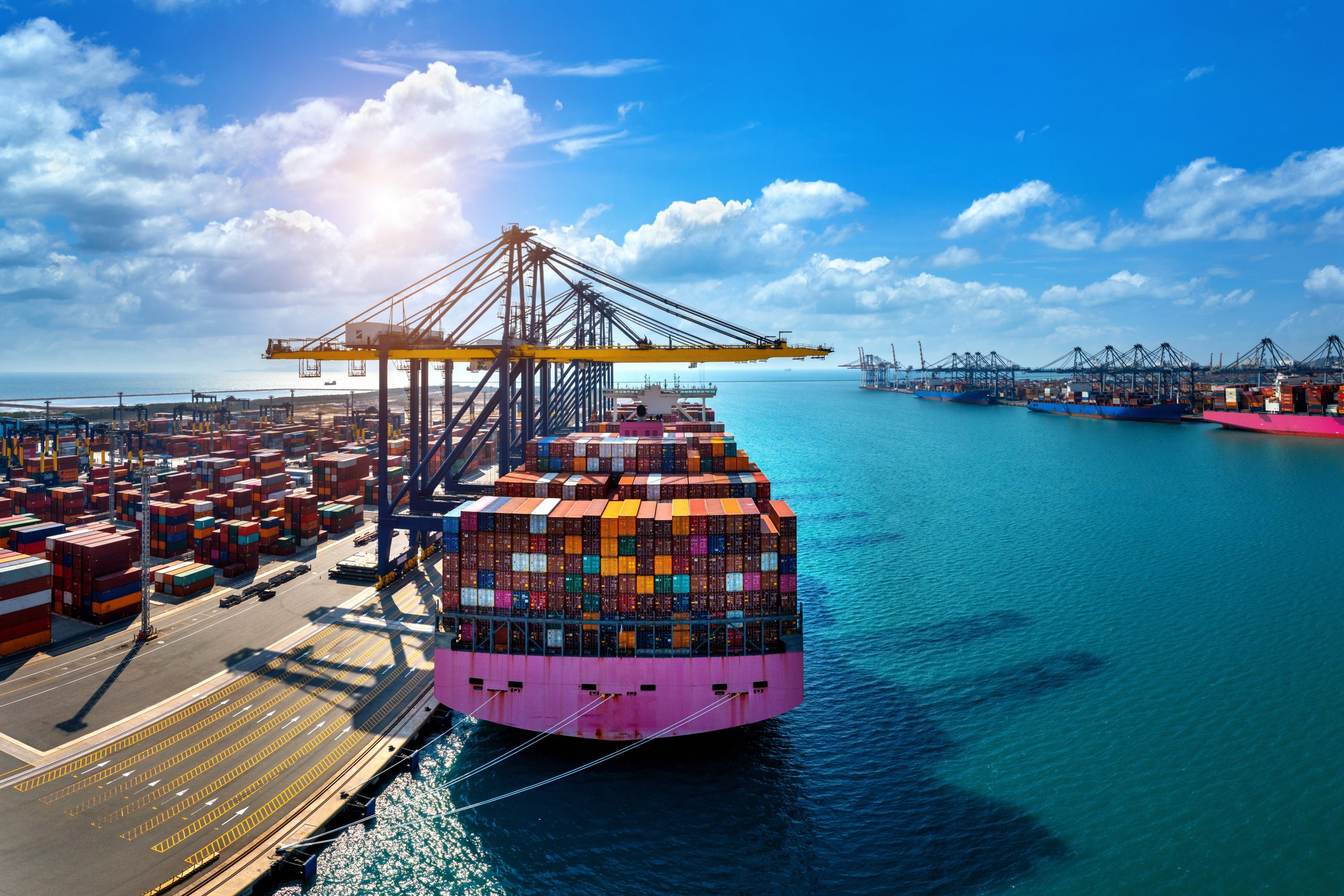Aerial view of cargo ship and cargo container in harbor - Vantree - Vantree Shipping port with multicolored stacked containers, a large yellow gantry crane, and blue sea under a sunny sky.