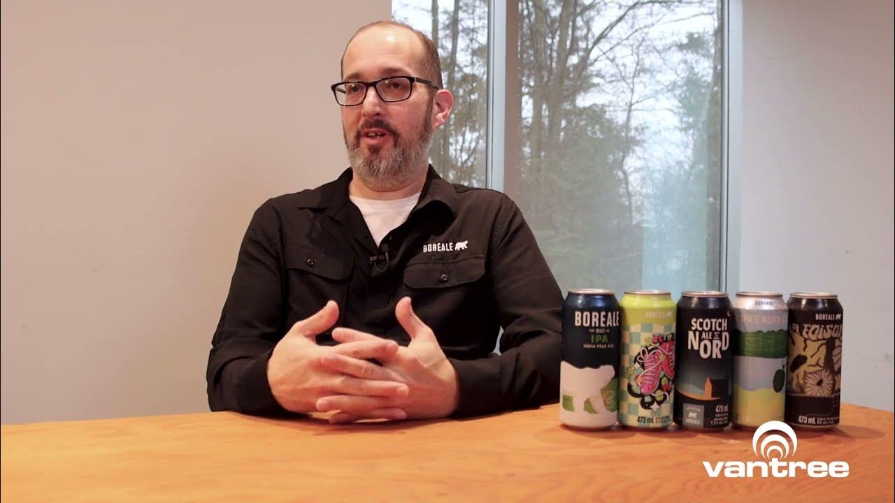 Man with glasses and beard sits at a wooden table during an interview, with several beer cans lined up on the right and a Vantree logo visible in the corner.
