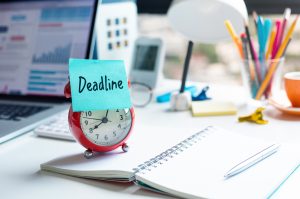 Deadline and work concepts with text on alarm clock on desk - Vantree - Vantree Red alarm clock with a blue 'Deadline' sticky note on a cluttered desk near a laptop and notebooks, signaling a due date in work.