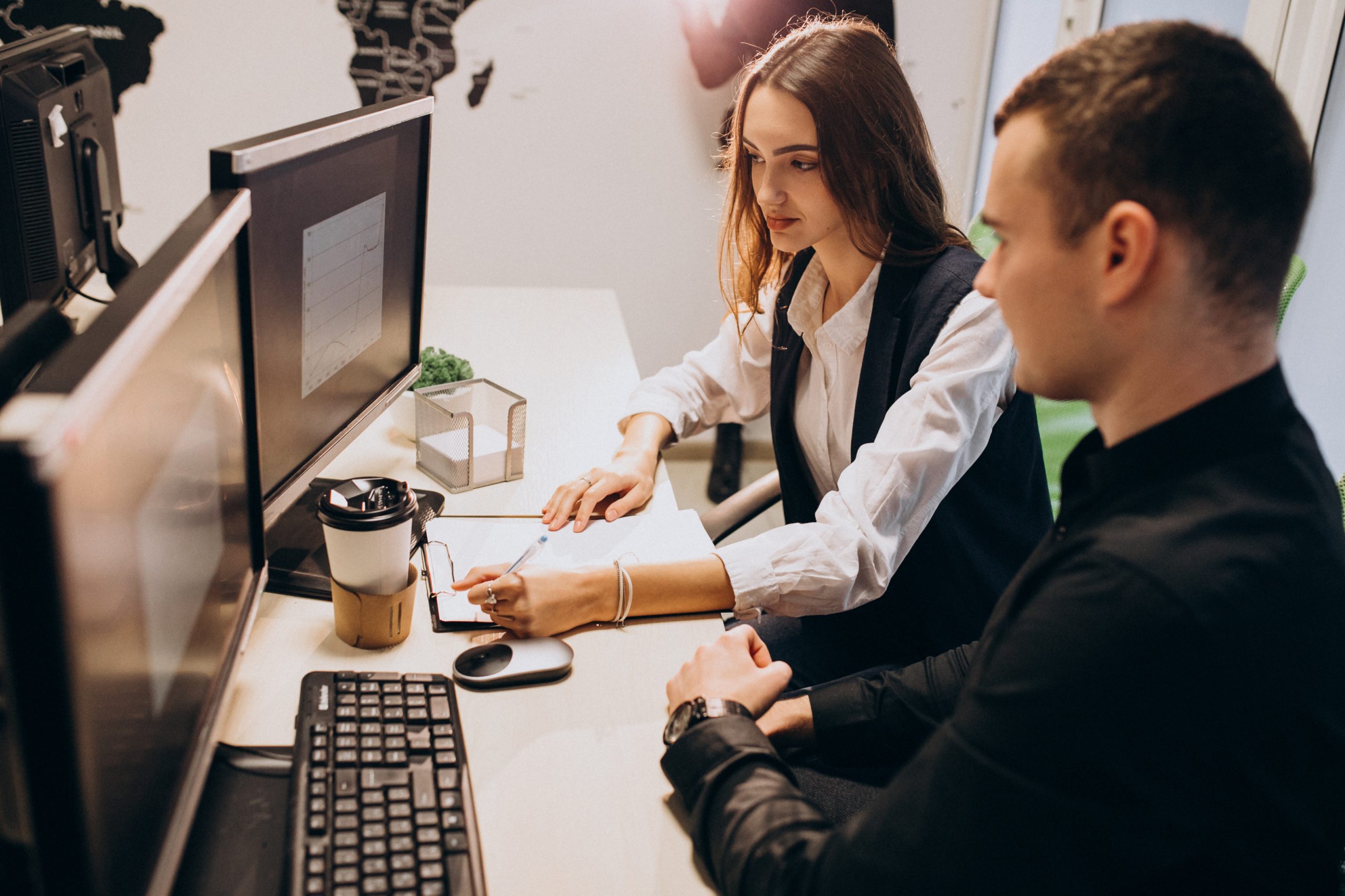 Workers at an IT company working on a computer - Vantree - Vantree Two coworkers work at a shared desk with computer monitors, keyboard, and papers in an office environment, coffee cup nearby.