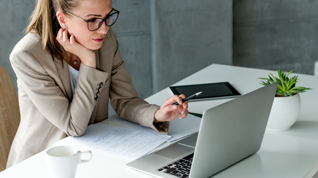 Businesswoman with glasses reviews documents at a laptop in a bright office setting on a white desk, pen in hand.