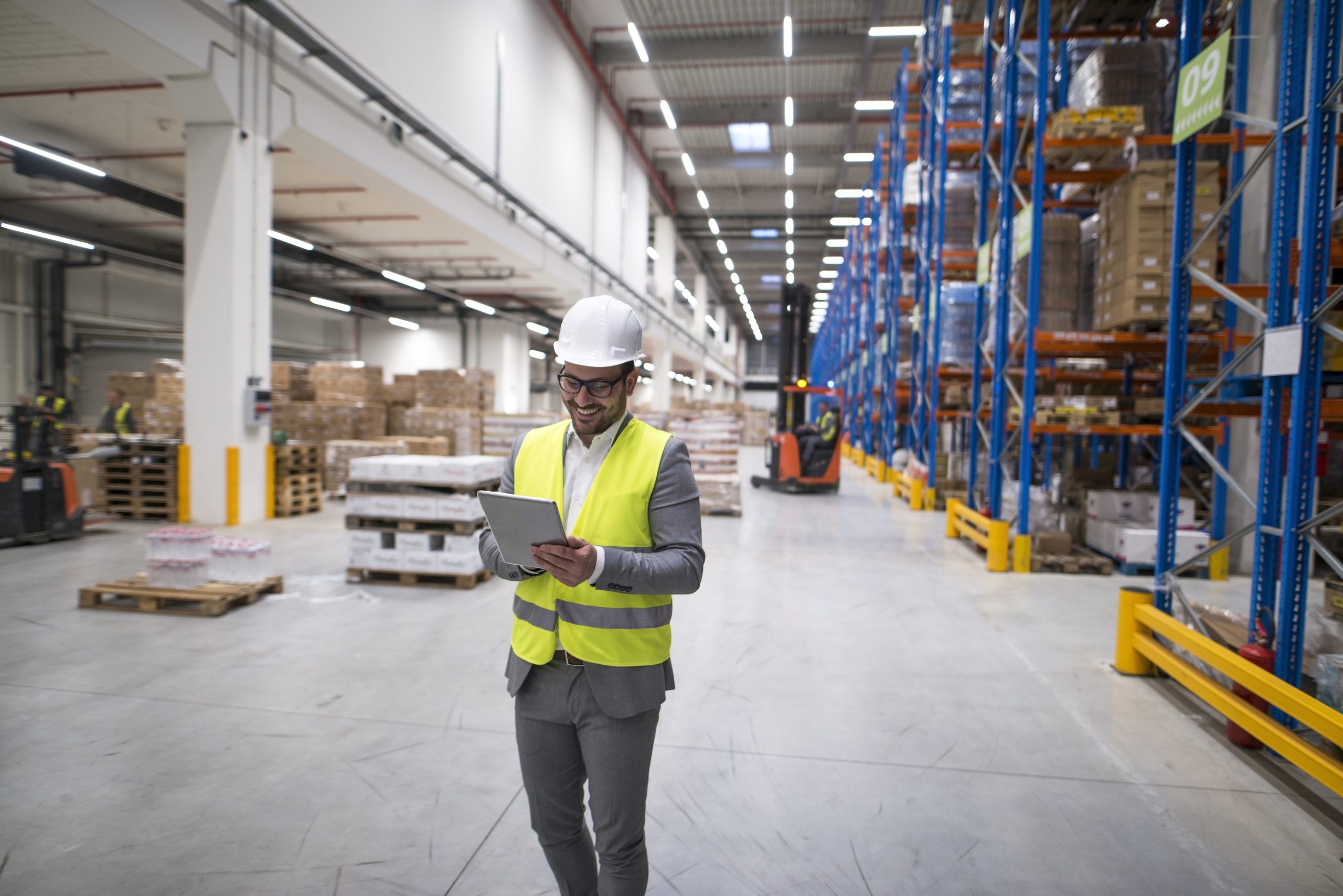 Man in a white hard hat and neon vest stands in a warehouse, looking at a tablet.